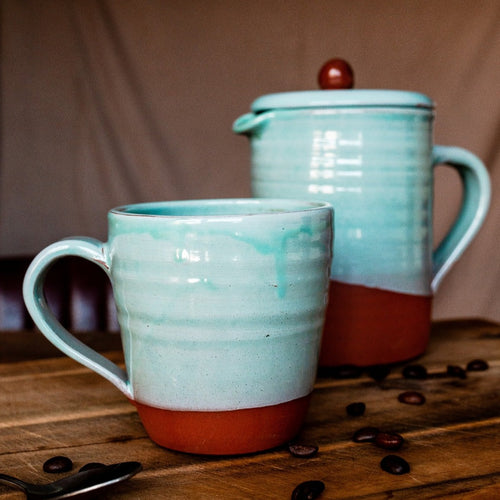 Large turquoise mug and cafetiere on a wooden board with coffee beans scattered around.
