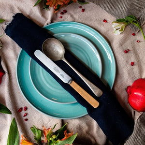 Turquoise side plate sitting on a turquoise dinner plate on a dark wooden table with a navy napkin soupspoon and butter knife.