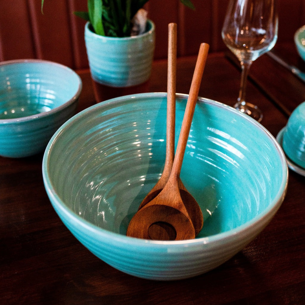 Large turquoise bowl with 2 wooden serving spoons inside. A turquoise bowl, vase and wine glass in the background.