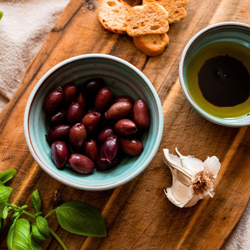 Medium turquoise bowl with black olives and a small turquoise bowl with olive oil and balsamic on a chopping board with mini bread slices and basil.