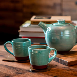 2 turquoise mugs and a turquoise teapot sitting on a wooden table with books in the background.